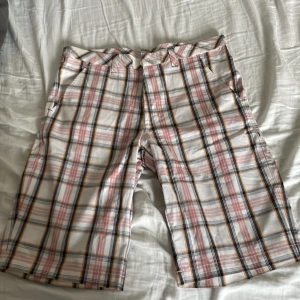 Rutiga shorts från Manlio, storlek 32 - Snygga rutiga shorts från Manlio i vitt med rosa, svarta och gula ränder. Klassisk passform med fickor och bälteshällor, perfekta för sommardagar. Tunt och lätt material som känns skönt mot huden. Styla med en enkel t-shirt för en chill look.