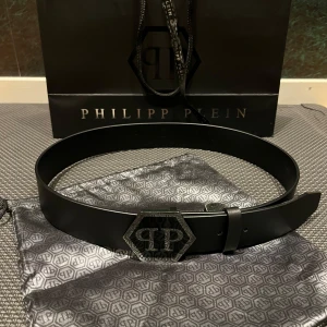 Svart skärp från Philipp Plein - Snyggt svart skärp från Philipp Plein i äkta skinn med en stor, sexkantig metallspänne med PP-logga. Skärpet är stilrent och har exklusiv känsla, perfekt för dig som gillar statement-accessoarer. Tillverkat i Italien och levereras med originalpåse.