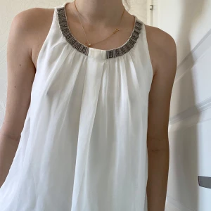 Vit ärmlös blus med pärldetaljer - Säljer en luftig vit blus med snyggt fall och plissering framtill. Halsringningen är dekorerad med silvriga pärldetaljer som ger en cool touch. Blusen är ärmlös och har en liten knappöppning i nacken. Perfekt för dig som gillar stilrena och trendiga plagg.