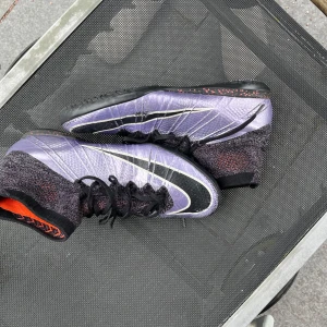 Lila Nike fotbollsskor strl 43 / Nike Superfly Indoor Football shoes - Säljer ett par lila Nike Superfly fotbollsskor (Elite) i storlek 43 med snygg lila och svart design, högt stickat skaft och orange detaljer. Skorna har snörning och svart sula med röda stänk. Perfekta för dig som vill sticka ut på planen.