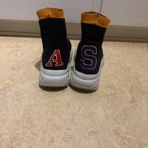Acne studio Svarta sock-sneakers  - Unika svarta sock-sneakers med vit chunky sula och orange ribbad kant upptill. Bak på hälen finns stora bokstäver i rött och lila. Skorna är i stickad syntet och har en modern, sportig vibe som sticker ut.