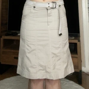 Beige cargokjol med bälte - Snygg beige kortkjol från Isolde med vita sömmar och tillhörande randigt bälte. Kjolen har praktiska fickor både fram och bak samt dragkedja framtill. Perfekt för dig som gillar en clean och enkel stil med en y2k touch.