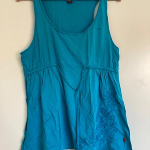 Turkos linnetopp från QS by s.Oliver - Säljer en turkos ärmlös linnetopp från QS by s.Oliver i bomull. Toppen har broderade blommor nertill på ena sidan och en avslappnad passform med lätt rynkade sömmar under bysten. Perfekt för varma dagar och enkel att styla med jeans eller shorts.