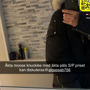 Svart Moose Knuckles dunjacka med päls - Säljer en svart dunjacka från Moose Knuckles med äkta päls på huvan. Jackan har snygga metalldetaljer, dragkedjor och knappar, samt en patch på ärmen. Perfekt för kalla dagar och riktigt stilren look.