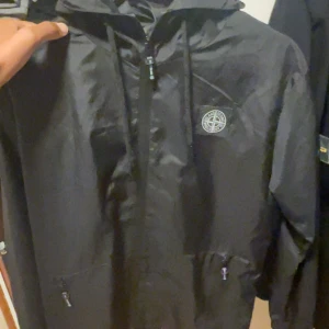 vindjacka stone island - Svart vindjacka från Stone Island med klassisk kompasspatch på ärmen och logga på bröstet. Jackan har huva, dragkedja framtill, snörning och två fickor med dragkedja. Tillverkad i 100% bomull, perfekt för dig som gillar streetwear och stilren design.