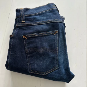 Nudie jeans - Snygga mörkblå jeans från Nudie Jeans med klassiska kontrastsömmar och vågig brodyr på bakfickan. Jeansen har en rak passform och är tillverkade i mjukt denimtyg. Perfekta för en stilren och avslappnad look.