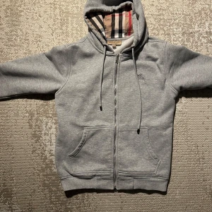 Grå hoodie från Burberry  - Snygg grå hoodie från Burberry med klassiskt rutigt foder i huvan. Tröjan har hel dragkedja, snörning i huvan och två stora fickor framtill. Perfekt för dig som gillar stilrena och exklusiva plagg. Kom hjärna med bud