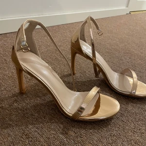 Guldiga sandaler med klack - Snygga sandaler i metallic guld med smala remmar och öppen tå. Klackarna är höga och ger en elegant look. Perfekta för dig som vill sticka ut med stilrena och trendiga skor. Remmarna går runt vristen och ger en säker passform.