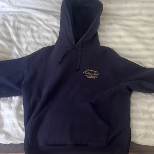 Mörkblå hoodie från Axel Arigato - Säljer en mörkblå hoodie från Axel Arigato med broderad text på bröstet. Tröjan har huva med snörning, stor känguruficka och är tillverkad i 100% bomull. Perfekt för dig som gillar stilrena och bekväma plagg med premiumkänsla. Skriv Järna om ni är intresserade