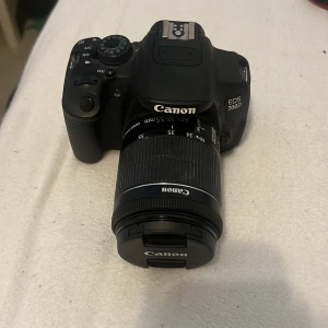 Cannon EOS 700D kamera  - Bilderna visar en Canon EOS 700D systemkamera med ett 18-55mm objektiv. Kameran är svart och har flera manuella inställningar, perfekt för dig som vill ta din fotografering till nästa nivå. Passar både nybörjare och dig som vill utvecklas inom foto.