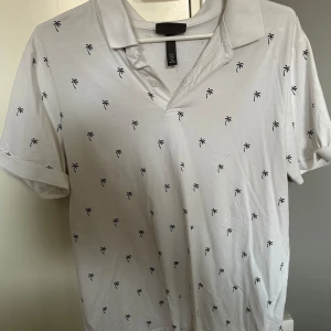 Vit piké med palmmönster från H&M - Vit pikétröja från H&M med små svarta palmer över hela tröjan. Klassisk krage och korta ärmar, perfekt för en chill sommarlook. Tillverkad i mjuk bomull som känns skön mot huden.