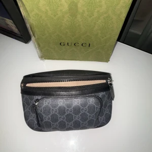 Svart Gucci axelväska med GG-mönster - Säljer en svart axelväska från Gucci med klassiskt GG-mönster i grått. Väskan har två dragkedjor, en större och en mindre ficka framtill. Tillverkad i skinn med struktur och detaljer i svart. Perfekt för dig som vill ha en stilren och lyxig accessoar.