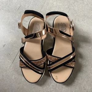 Glittriga sandaler i guld och svart - Snygga sandaler från Petite Sofie Schnoor i storlek 37. De har svarta och guldiga remmar, varav några är glittriga, och en bekväm platt sula. Remmarna är i syntetmaterial och sandalerna har spänne vid ankeln. Perfekta för att lyfta din sommarstil.