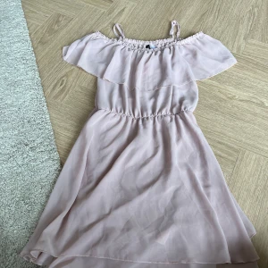 Ljusrosa offshoulder klänning Divided - Supersöt ljusrosa kortklänning från Divided med offshoulder och volang upptill. Klänningen har smala axelband, resår i midjan och är tillverkad i ett lätt, flowy material som ger en romantisk vibe. Perfekt för varma dagar och somriga tillfällen.
