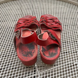 Röda svenska träskosandaler i skinn - Snygga röda sandaler med flätade remmar i skinn och justerbart spänne. Tillverkade i Sverige med klassisk träskosula och öppen tå. Perfekta för sommarens alla äventyr och ger en retrovibe till din outfit. Använda syns vid hälen och fotsulorna, se bild, pris kan diskuteras!