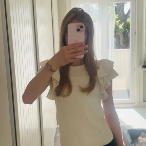 Ribbad vit topp med volangärmar - Ribbad topp i vitt med volangärmar som ger en extra touch. Ärmlös modell med rund halsringning och mjukt, stretchigt tyg. Passar perfekt till både jeans och kjol. 