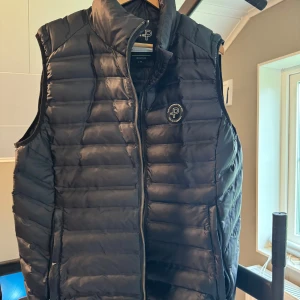 Svart dunväst från Pelle P - Snygg svart dunväst från Pelle P med quiltad design och dragkedja framtill. Västen har hög krage och ett broderat Pelle P-märke på bröstet. Perfekt att slänga över en hoodie eller tröja när det är kyligt ute. Ny pris ca 1500 och helt o använd men säljer endast för 299 priset är diskuterbart!