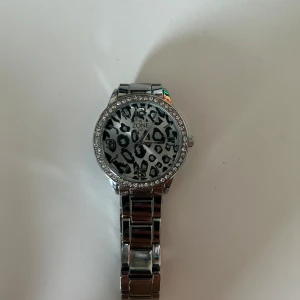 Silverfärgad klocka med leopardmönster - Stilren klocka från IN ZONE med glittrande stenar runt urtavlan och ett coolt svartvitt leopardmönster. Klockan har en länkad metallrem i silver och tydliga siffror på urtavlan. Perfekt accessoar för dig som vill sticka ut. Oklart om klockan fungerar för den inte använts på länge(batteri kan behövas bytas). 
