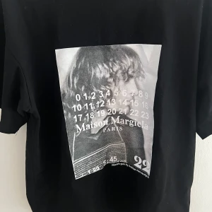 Maison Margiela svart t-shirt med print - Svart t-shirt från Maison Margiela med ikoniskt grafiskt print framtill i svartvitt. Klassisk passform, rund hals och korta ärmar. Tillverkad i mjuk bomull och har de typiska vita sömmarna bak i nacken. Perfekt för dig som gillar statement-plagg.