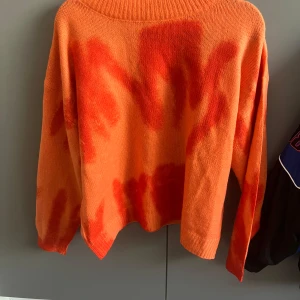 Orange stickad tröja med röd tie-dye - Säljer en oversized stickad tröja i orange med röda tie-dye detaljer. Tröjan har lång ärm, bred ribbad krage och avslappnad passform. Perfekt för dig som gillar färg och vill sticka ut lite extra. Lappen är avklippt så vet ej storlek men skulle tippa på S och påbörjad Medium då den är lite oversized.