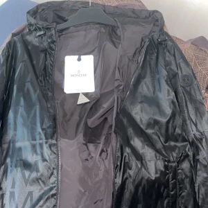Svart Moncler vindjacka, limited edition - Svart Limited edition vindjacka från Moncler som är väldigt sällsynt med unikt mönster i tyget och snygg svart moncler märke på ärmen. Jackan har huva med snörning, dragkedja framtill och är tillverkad i glansig polyester. Perfekt för dig som vill ha en exklusiv look, Jackan går även att byta håll på för att få den hel svart ifall du vill ha en stilren look. Jackan är använd 1 gång och är tyvärr tvungen o sälja den då jag är i behov av pengar. Den är storlek S men passar även M, 