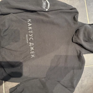 Svart Cactus Jack hoodie Travis Scott - Svart hoodie från Cactus Jack x Travis Scott med rysk text och tryck på bröstet samt stor grafisk design på ryggen. Klassisk känguruficka och huva. Perfekt för dig som gillar streetwear och unika prints.