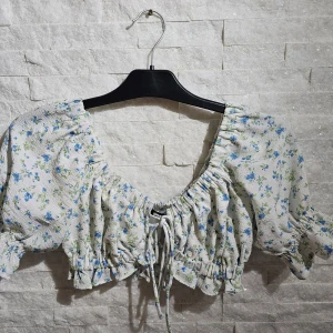 Blommig croppad blus från Ginatricot - Supersöt croppad blus från Ginatricot med blått och grönt blommönster på vit botten. Toppen har puffiga trekvartsärmar med volangkant, v-ringning och snörning framtill. Perfekt för vår och sommar när du vill ha en romantisk och trendig look.