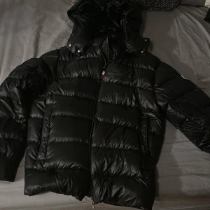 Svart Moncler dunjacka med huva - Säljer en svart dunjacka från Moncler med glansig finish och stor huva. Jackan har klassisk pufferstil, dragkedja framtill och Moncler-logga på ärmen. Perfekt för kalla dagar när du vill hålla stilen och värmen.