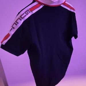 Svart - Designer Piké - Snygg svart pikétröja med korta ärmar, vit och röd krage samt matchande ränder och logga längs axlarna. Tröjan har knappar vid halsen och är gjord i mjuk bomull. Perfekt för dig som vill sticka ut med en elegant och trendig look. Tröjan är i nyskick, och priset går att diskutera vid snabb, smidig affär. Skriv i DM om ni vill ha detaljer om tröjan såsom märket🎶