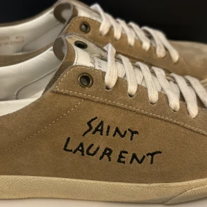 Saint Laurent mocka sneakers - Snygga sneakers från Saint Laurent i beige mocka med vit sula och vita snören. Handritad svart logga på sidan ger en cool och unik vibe. Perfekta för dig som gillar stilrena och exklusiva skor. Kom med bud! 