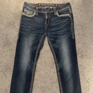 Rock Revival jeans med broderade fickor - Snygga mörkblå jeans från Rock Revival med kontrastsömmar och broderade bakfickor. Jeansen har slitningar och tvättade detaljer framtill och baktill, samt klassisk femficksdesign. Perfekta för dig som gillar en unik och detaljerad look. Skiriv om ni har ytterligare frågor!