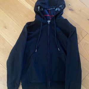 Burberry hoodie  - Svart hoodie från Burberry med klassiskt rutigt foder i huvan och broderad logga på bröstet. Tröjan har dragkedja framtill, snörning i huvan och stora fickor. Tillverkad i mjuk bomull och har en clean, stilren look.