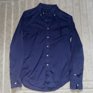 Blå skjorta från Ralph Lauren slim fit - Snygg blå skjorta från Ralph Lauren i slim fit-modell. Tillverkad i lätt bomullstwill och har klassisk knappslå framtill samt vit knappdetalj. Ikoniska röda Polo-loggan broderad på bröstet. Perfekt för dig som gillar stilrena och tidlösa plagg.