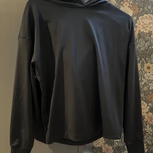 Svart träningshoodie med huva - En svart hoddie i träningsmaterial, detta är en kortare model som slutar ungefär vid höften. Materialet är lent och ganska tunt så perfekt när det är varmt och lite kyligt!