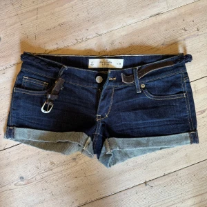 Mörkblå jeansshorts Abercrombie & Fitch - Snygga mörkblå jeansshorts från Abercrombie & Fitch med uppvikta benslut, klassiska fickor och flätat bälte i midjan. Shortsen har kontrastsömmar och broderade detaljer på bakfickorna. Perfekta för varma dagar och en avslappnad look.