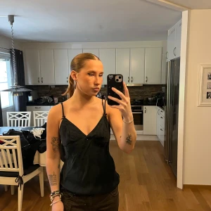 Svart linnetopp med rosetter vintage från Italien  - Svart linnetopp med smala axelband och små söta rosetter vid axelbanden. Toppen har v-ringning och är i ett lätt glansigt material som känns lyxigt. Vintage från Italien så har inga märkeslappar men uppskattats i storlek S.