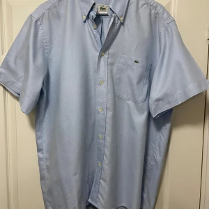 Ljusblå kortärmad skjorta Lacoste - Klassisk ljusblå skjorta från Lacoste med klassisk button-down krage och korta ärmar. Skjortan har en bröstficka med den ikoniska krokodilloggan broderad. Tillverkad i bomull och stängs med vita knappar framtill. Kvalitétstyg.