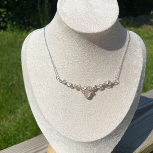 Handgjort halsband med rosa sten (rosenkvarts) - Unikt halsband i silverfärgad metall med en ljusrosa hjärtformad sten i mitten, infattad i snirkliga detaljer. Kedjan är tunn och justerbar med karbinlås. Perfekt för dig som gillar boho eller romantisk stil och vill ha något som sticker ut.
