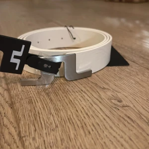 Vitt skärp från J.Lindeberg med silverfärgat spänne - Snyggt vitt bälte från J.Lindeberg med ett stilrent, matt silverfärgat spänne i metall. Bältet är gjort i äkta skinn och har en modern design som passar perfekt till jeans eller kostymbyxor. Längd 110 cm. 10/10 skick, tags kvar. priset är diskuterbart,😇 Nypris 599kr. hör av dig vid frågor👌