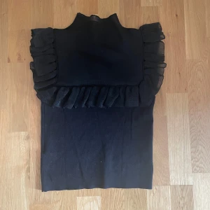 Svart volang topp med hög hals - Supersnygg svart topp med hög hals och volangdetaljer över axlar och bröst. Toppen är ärmlös och väldigt trendig. Köparen står för frakt 