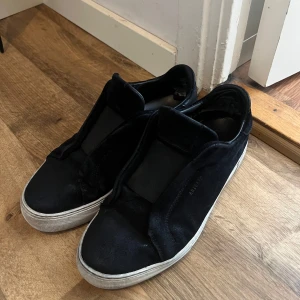 Svarta sneakers från Axel Arigato - Stilrena svarta sneakers från Axel Arigato i mocka med vit platt sula. Skorna har minimalistisk design utan synlig snörning och en diskret logga på sidan. Perfekta för dig som gillar en clean och modern look. Är i bra skick 