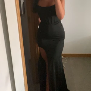Svart balklänning med slits - Svart balklänning med tight men stretchig passform och slits. Korsetliknande design som formar kroppen fint. Endast använd en gång. Skriv ifall ni vill ha fler bilder