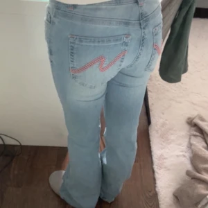 Ljusblå bootcut jeans från Nelly - Säljer ett par ljusblå jeans från Nelly med bootcut-modell. Jeansen har klassisk femficksdesign, rosa broderad detalj på bakfickorna och låg midja. Tillverkade i mjukt denimtyg med en skön passform som sitter snyggt över höfterna och släpper ut vid benen.