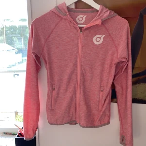 Rosa sportig vindjacka med huva - Säljer en rosa vindjacka från Kappahl i sportig stil. Jackan har huva, hel dragkedja framtill och två fickor med dragkedja. Materialet är lätt och syntetiskt, perfekt för utomhusaktiviteter. Långärmad modell med tumhål i ärmsluten och diskret mönster.