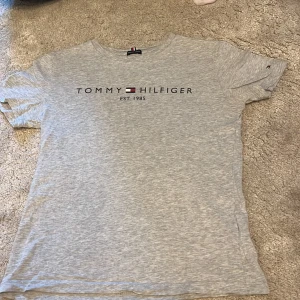 Grå t-shirt från Tommy Hilfiger - Snygg grå t-shirt från Tommy Hilfiger i ekologisk bomull. Klassisk modell med korta ärmar och rund hals. Framsidan har stilren logga och texten 'EST. 1985'. Perfekt basic-plagg med diskret flaggdetalj på ärmen.