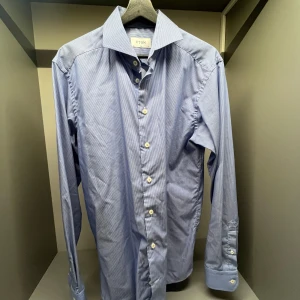 Blå randig skjorta från Eton slim fit - Snygg blå skjorta från Eton i slim fit med tunna vita ränder. Klassisk krage, långa ärmar och vita knappar framtill. Perfekt för dig som gillar stilrena och tidlösa plagg. Tillverkad i mjuk bomull för en skön känsla hela dagen.