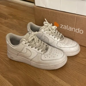 Vita Nike Air Force 1 sneakers - Klassiska helvita Nike Air Force 1 sneakers i skinn med perforerad tå, rund tå och snörning. Snygg och tidlös modell med låg profil och ikonisk Nike-logga på sidan och hälen. Perfekta för dig som gillar streetstyle och vill ha en clean look.