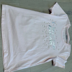 Ljusrosa t-shirt med pärldetaljer - Ljusrosa t-shirt från Cosmoda med texten 'I DON'T CARE ANYMORE' framtill, dekorerad med små pärlor. Klassisk rund hals och korta ärmar. Skön och mjuk i bomull, perfekt för en chill look.