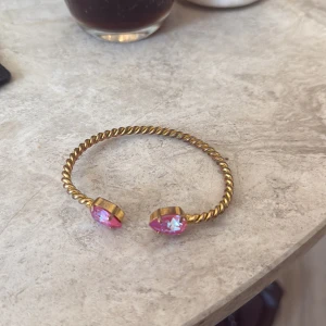 Caroline Svedbom armband med rosa stenar - Snyggt armband i guldtonad metall med tvinnad design. Öppna ändar pryds av två glittrande rosa stenar som ger en färgklick och lyxig känsla. Perfekt accessoar för att lyfta din outfit med lite extra bling.