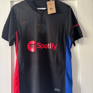 FC Barcelona Lamine Yamal Nike XS tröja - Snygg svart FC Barcelona fotbollströja från Nike med Lamine Yamal och nummer 19 på ryggen. Tröjan har röda och blå detaljer på sidorna, klubbmärke på bröstet och Spotify-logga framtill. Tillverkad i lätt och ventilerande material, perfekt för match eller träning.